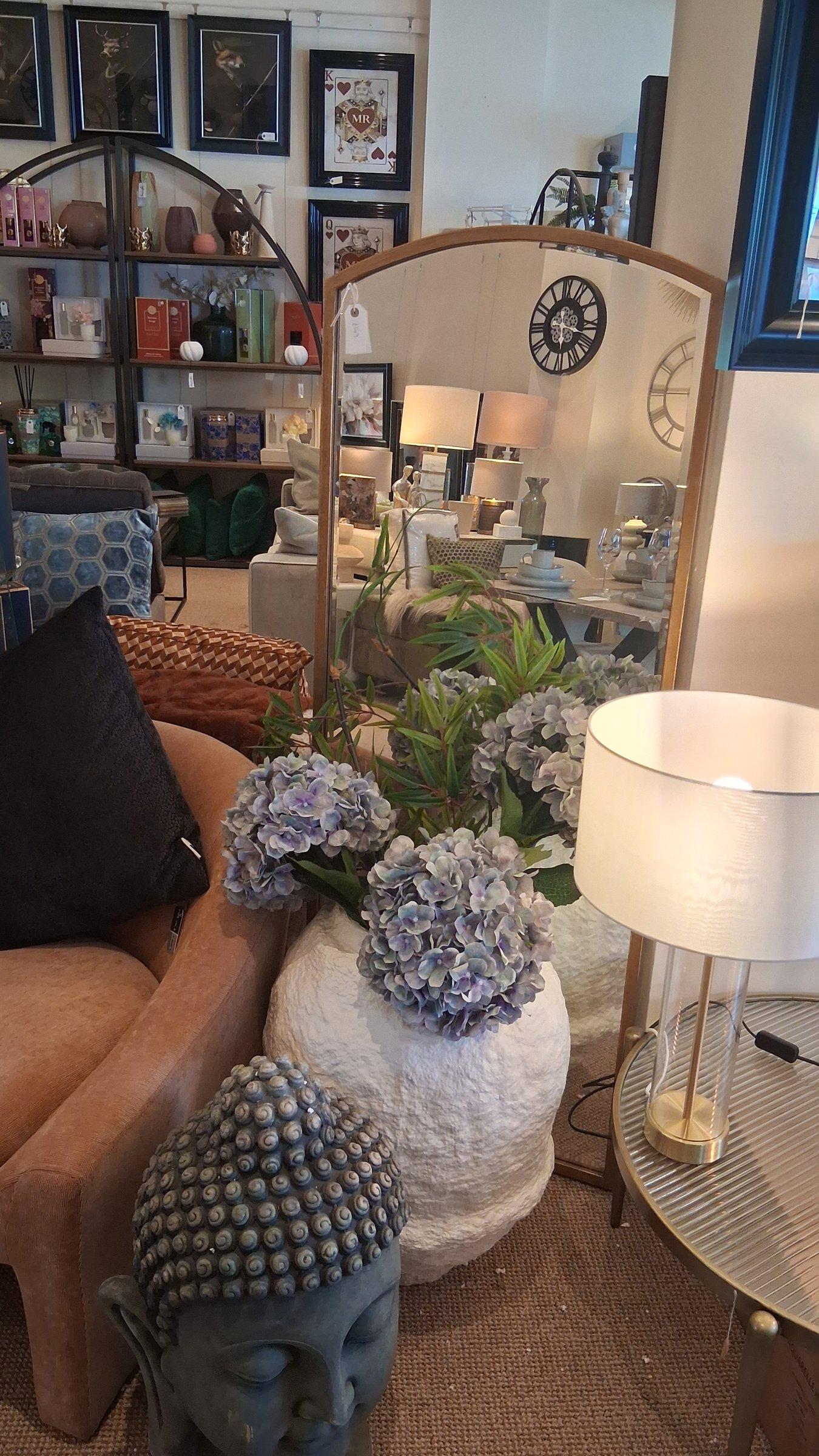 Tall arched mirror reflecting the showroom, with hydrangeas in a stone vase