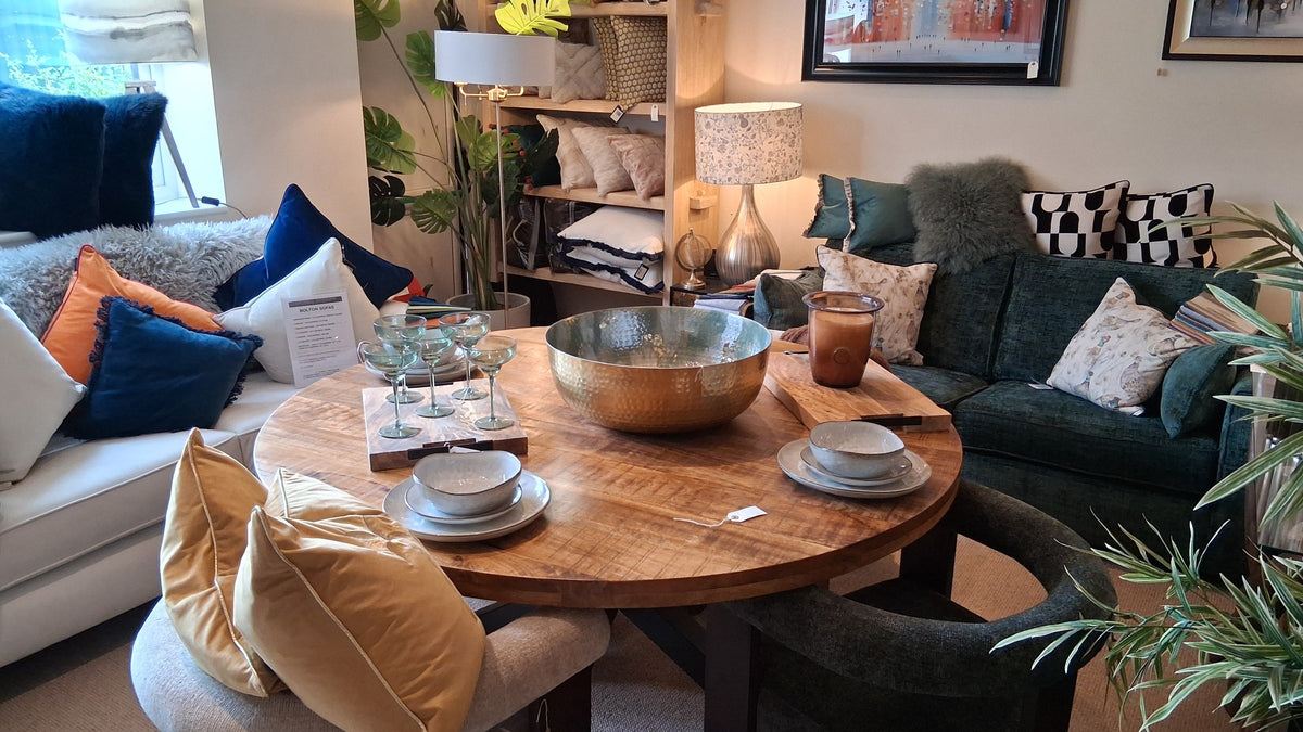 A round wooden dining table set with bowls and glasses beside a green sofa and cushions in the Canwell showroom